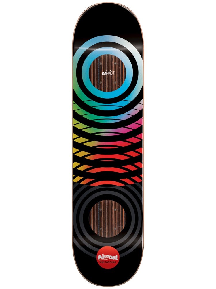 Almost Yuri Black Blur Impact 8.5 skateboard deck: Concentric rings in a spectrum gradient from blue through yellow to red fade to grey on a black deck, with two circular wood-grain windows showing the Impact construction. A red Almost logo sits near the tail.