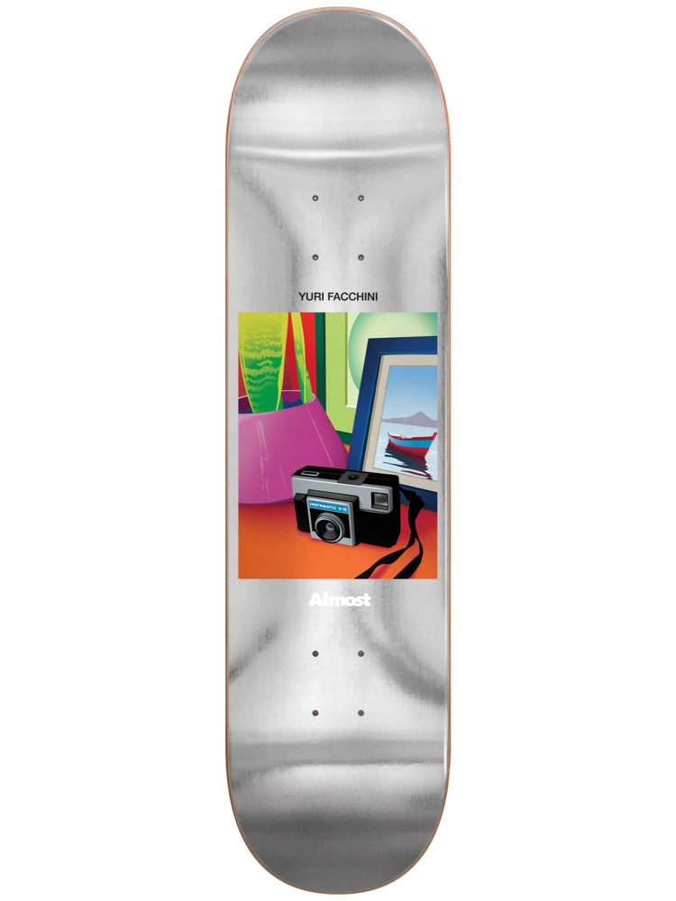 Almost Yuri Life Stills Impact Light 8.375 skateboard deck: Silver foil deck with a small framed illustration in the centre: a vintage instamatic camera on an orange table beside a pink bag, a green plant and a framed picture of a red boat on water.