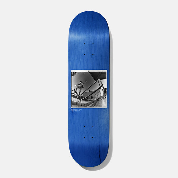 Baker Spanky BAKERVCA 8.25 skateboard deck: Black and white fisheye photograph of a skater mid-trick over a handrail beside a building, set in a white-bordered square on a blue-stained deck, with tiny caption text underneath.