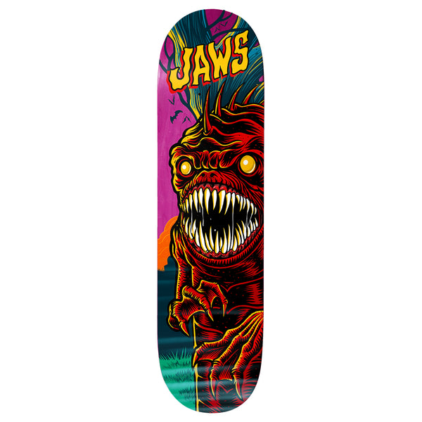Birdhouse Jaws Graveyard 8.475 skateboard deck: A red scaly monster with spiked head, glowing yellow eyes and a huge mouth of fangs climbs over a fence post, against a magenta and teal night graveyard scene. Yellow 'Jaws' lettering sits at the nose.