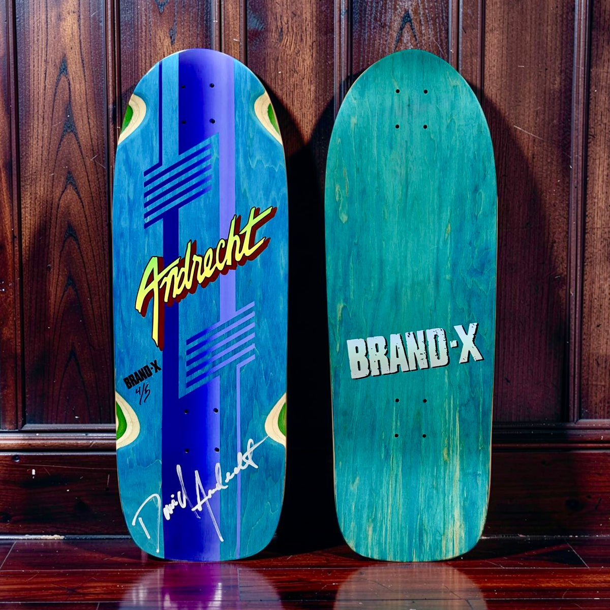 Brand-X Dave Andrecht skateboard deck: Old-school shaped deck stained blue, with a purple vertical stripe and stepped line motifs in the style of 1980s graphics, a bold yellow and red script signature name and a white handwritten autograph.