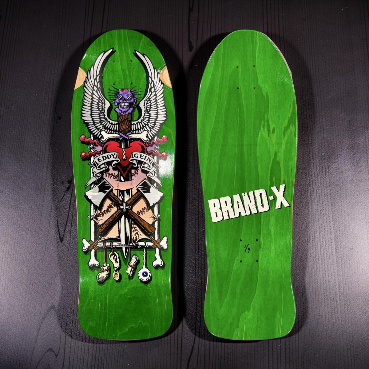 Brand-X Ed Gein Stain skateboard deck: Shaped old-school deck with a winged purple skull on a dagger through a stitched, broken red heart, a banner reading 'Eddy Gein', crossed axes and bones, and dangling ears and an eyeball. Green stain; underside has the brand logo.
