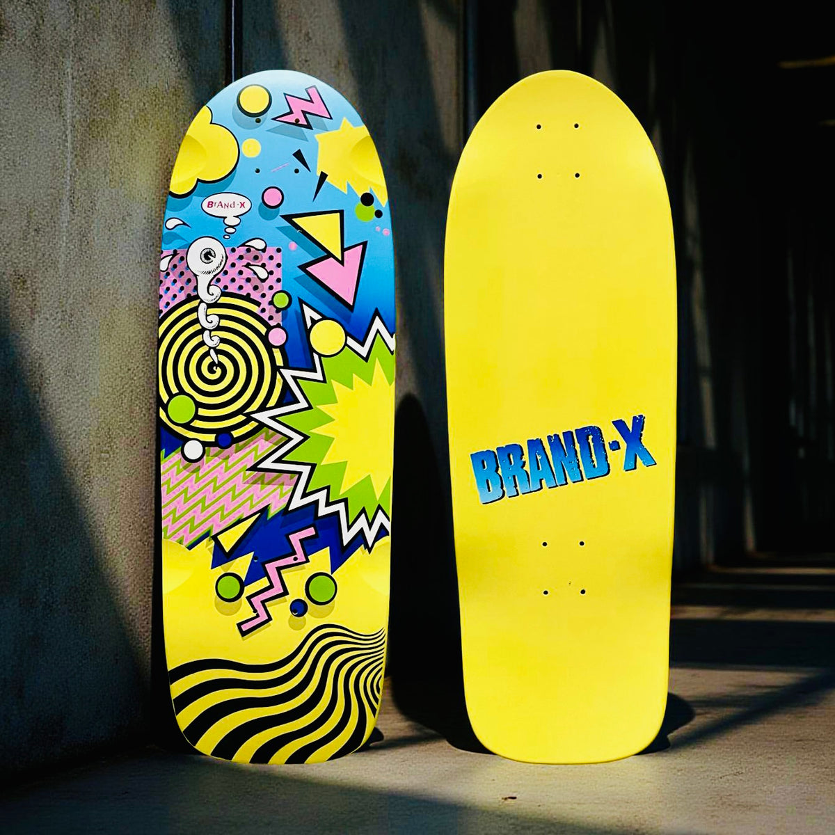 Brand-X Weirdo skateboard deck: Old-school shaped deck with a bright 1980s-style abstract design of spirals, zigzags, triangles, starbursts and dots on a blue and yellow background, with a small eyeball character. The other side of the photo shows plain yellow with 'Brand-X' in blue.