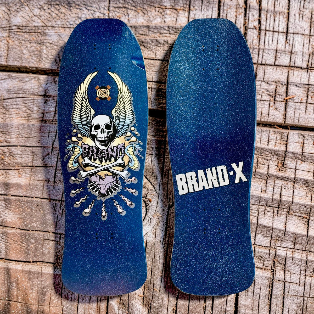 Brand-X X-Con skateboard deck: Winged skull above crossed bones and a heart, with 'Brand' lettering and snakes, in cream, grey and pink on a glittery navy blue old-school shaped deck. Underside shows 'BRAND-X' in white.