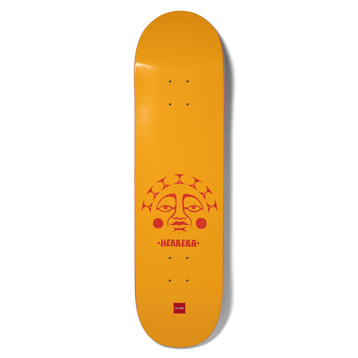 Chocolate Herrera Sunsign skateboard deck: A red stylised sun face with a rayed, patterned crown and round cheeks sits in the lower half of a plain orange-yellow deck, with the rider's surname beneath. A small red Chocolate logo is at the tail.