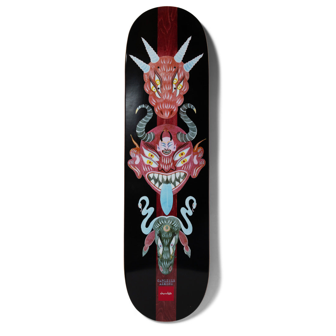 Chocolate Aikens Dog Perfume Mask skateboard deck: Vertical totem of three folk-art demon masks, a horned red dragon head, a fanged red demon with blue tongue and a green creature with snakes, over a dark red stripe on black. Small name text and Chocolate logo at the bottom.