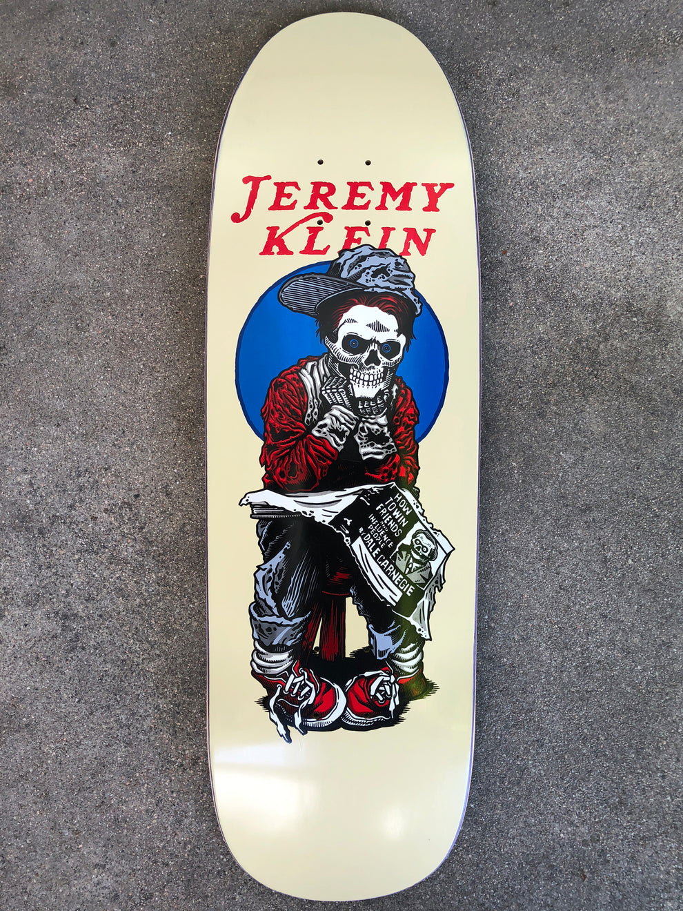 Hook-Ups Jk Industries Jeremy Klein black eye kid skull skateboard deck: Cream shaped deck showing a skeleton newsboy in a flat cap, red jumper and red trainers, seated on a stool reading a newspaper, in front of a blue circle. Rider name in red lettering above.