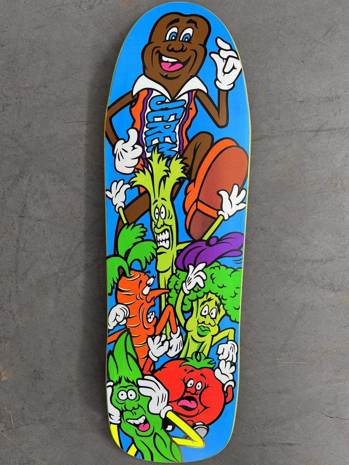 Hook-Ups Jk Industries Jeremy Klein Candy Bar skateboard deck: Cartoon chocolate bar character in an orange and blue jersey, strutting over a pile of panicking cartoon vegetables including celery, carrot, broccoli, tomato and lettuce, on a bright blue old-school shaped deck.
