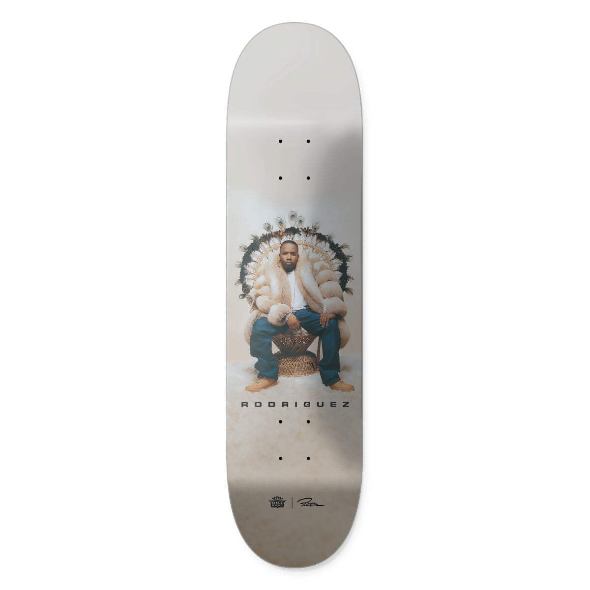 Primitive X Outkast Rodriguez Regal skateboard deck: Colour photograph of a man in a cream fur coat and jeans seated on a large wicker peacock chair edged with feathers, on a soft beige background. RODRIGUEZ in spaced capitals below, with Outkast and Primitive logos at the tail.