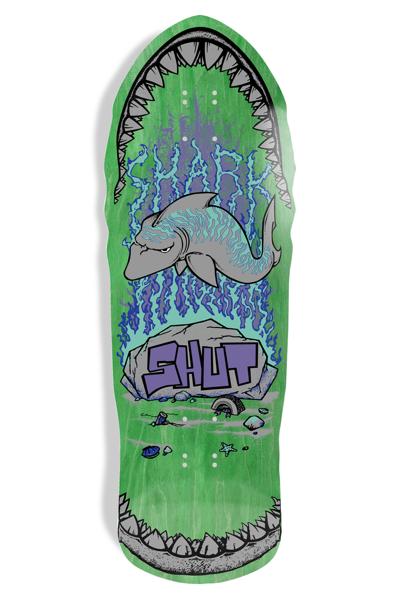 Shut 86 Shark EX7 (Green) skateboard deck: Old-school shaped deck on green stain showing a grey shark with a grumpy face swimming over a rock, with blue wavy water lines. The SHUT logo in purple graffiti letters sits on the rock, and shark jaws with teeth frame the nose and tail.