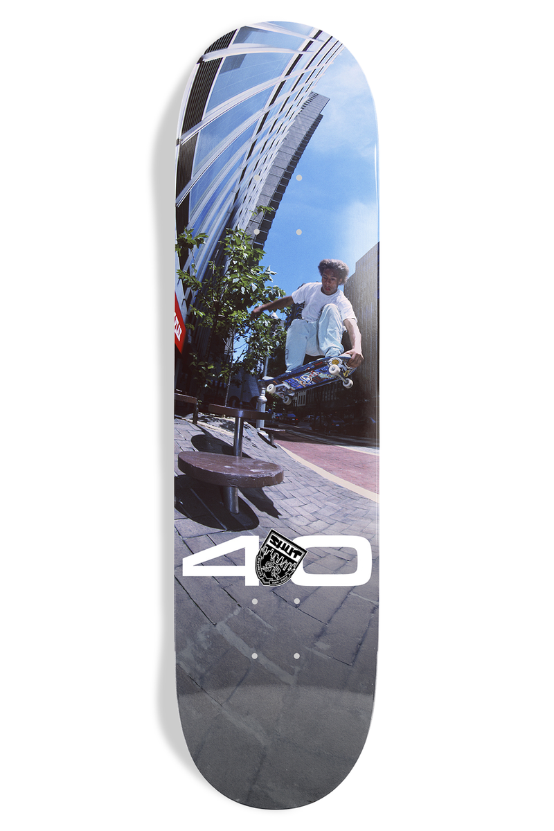Shut Bryce Kanights Sheffy EX7 skateboard deck: A colour photo of a skater in white clothes airing over a bench in front of a tall glass building, with a large white 40 and a small crest at the lower end.