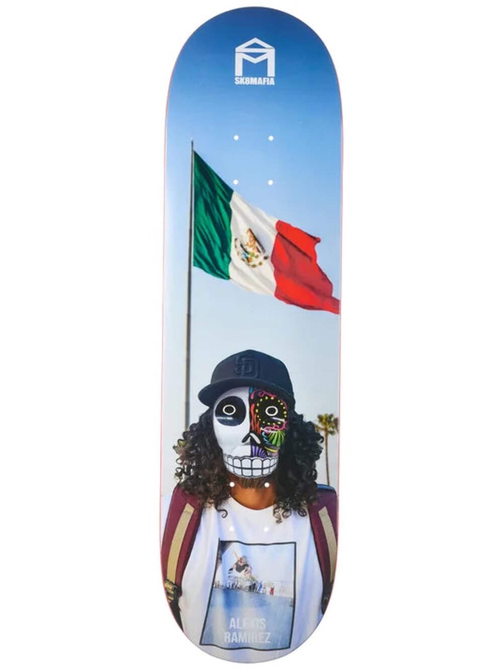Sk8Mafia Ramirez Dios skateboard deck: Colour photograph of a curly-haired person in a baseball cap and a half-skull Day of the Dead face mask, standing before a waving Mexican flag on a pole against a blue sky. The Sk8Mafia house logo is at the top.