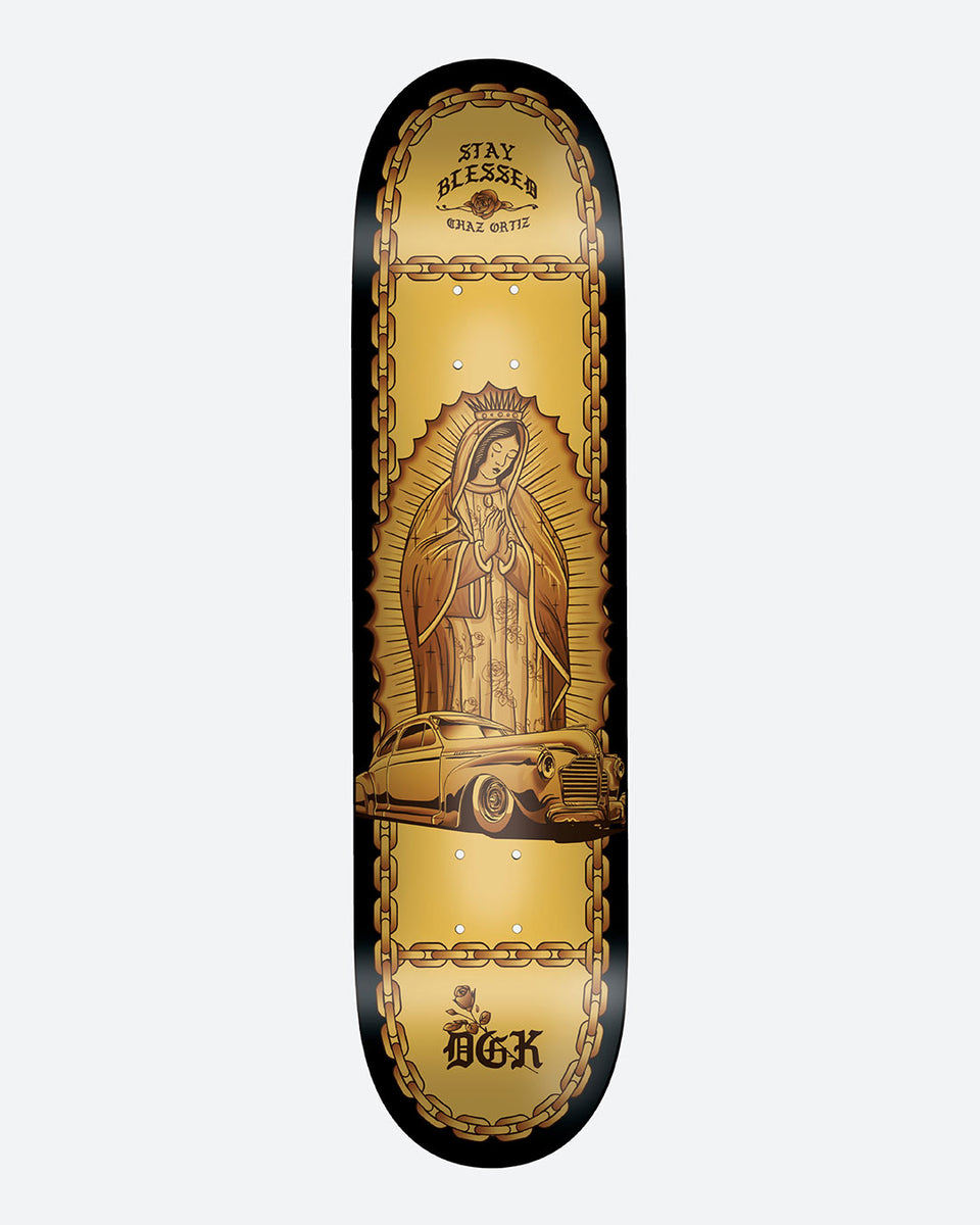 DGK Lo - Side Ortiz 8.1 skateboard deck: A praying Virgin of Guadalupe figure with a golden halo stands above a gold lowrider car, inside a chain-link border on a gold and black deck. Blackletter Stay Blessed banner at the top and DGK logo with a rose at the tail.