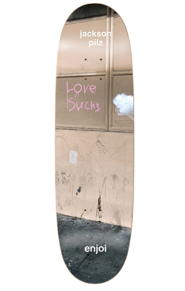 Enjoi Pilz Tweaker R7 8.25 & 9.125 skateboard deck: Photograph of a dirty street wall and pavement with pink graffiti reading 'Love Sucks' on a boarded window. Rider's name at the top and brand name at the tail, in white text on a shaped deck.