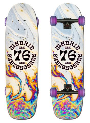 Madrid Grub 29.25 skateboard deck: A circular black 'Madrid Skateboards Est 76 USA' badge over a swirling oil-slick marbled pattern, white and pale grey at the top turning to rainbow at the bottom. Shown as a complete cruiser with purple wheels beside the bare deck.
