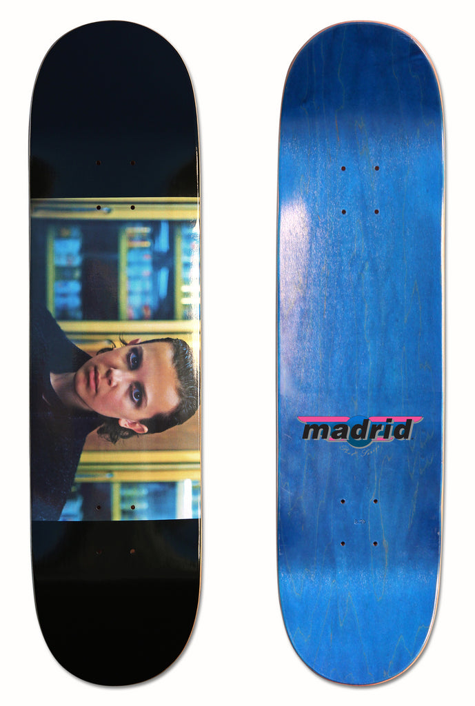 Madrid X Stranger Things - B*tchin Eleven 8 skateboard deck: Cinematic still of a young woman with short dark hair and heavy eye makeup, in dark clothes in front of shelves, as a wide horizontal photo band on a black deck. The blue underside carries the Madrid logo.