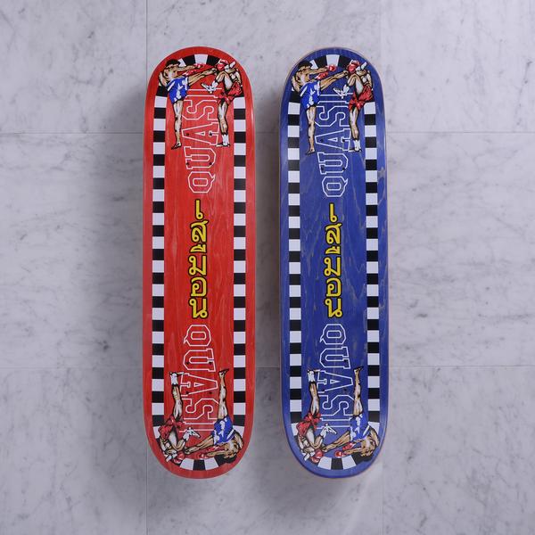 Quasi K.O. Red skateboard deck: Red-stained deck bordered with a black and white chequered edge, showing two Muay Thai fighters mid high kick at each end, with Quasi in outlined letters and yellow Thai script down the middle. Blue version shown beside it.