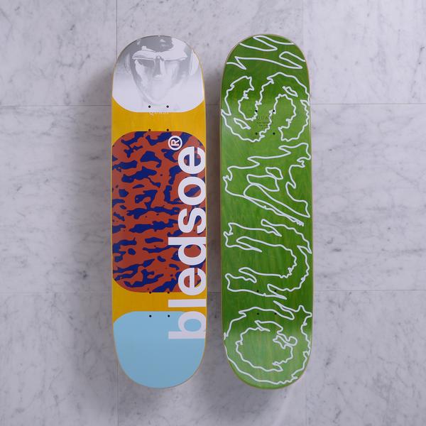 Quasi Metalhead Blue skateboard deck: Two decks shown. The top has a grey monochrome face at the nose, a yellow field with an orange and blue leopard-style blotch panel, and a light blue tail, with large white vertical lettering. The bottom is green with white scribbled outline lettering.