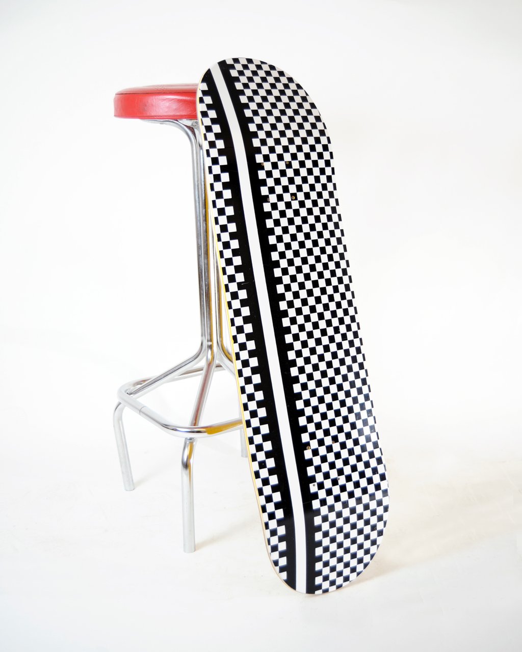 Skateboard Cafe Check Stripe Black/White skateboard deck: Black and white chequerboard pattern covering the whole deck, with a solid white stripe and black stripe running down the length of it. Shown leaning against a bar stool.