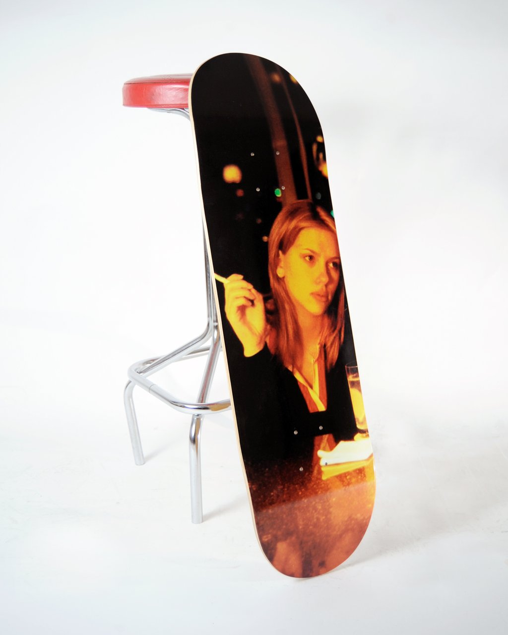 Skateboard Cafe LIT Charlotte skateboard deck: Warm-toned photograph of a woman with shoulder-length hair holding a cigarette, seated at a bar with a drink, with blurred amber lights behind her. Deck is photographed propped against a bar stool.