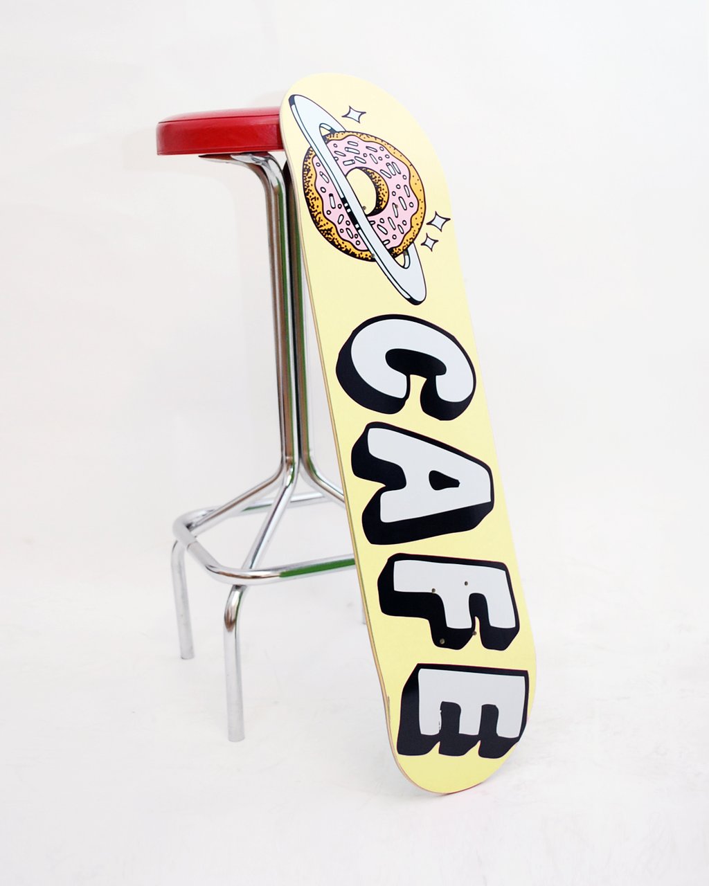 Skateboard Cafe Planet Donut Banana skateboard deck: Pink-iced sprinkled donut ringed like a planet, with sparkles, above chunky white Cafe lettering with black shadows on a pale yellow deck.
