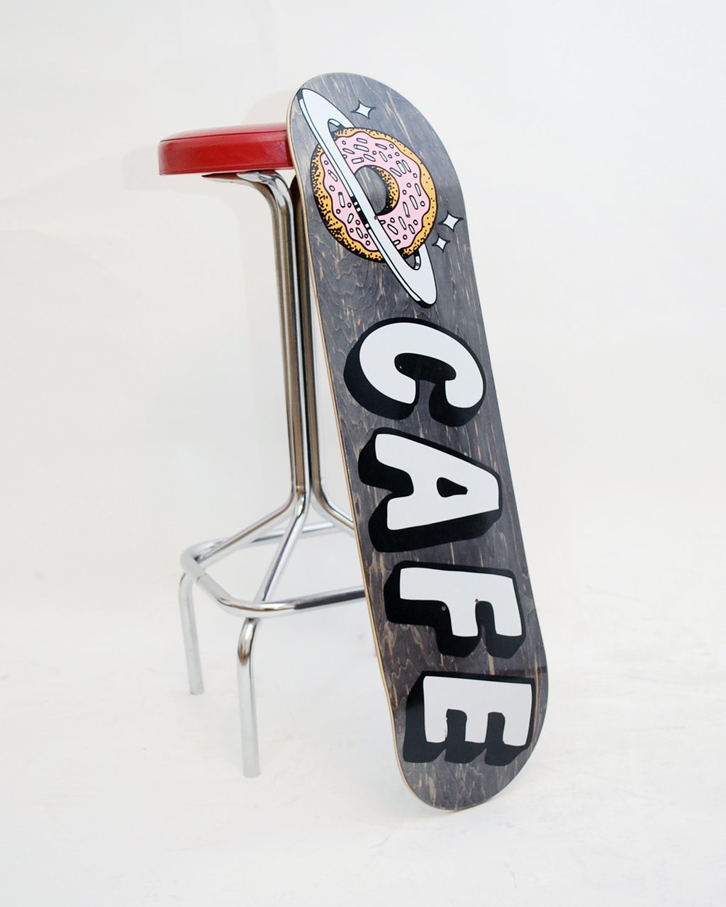 Skateboard Cafe Planet Donut Black skateboard deck: Dark grey stained deck with a pink iced doughnut ringed like a planet with stars at the nose, above large white block letters spelling CAFE with black shadows.