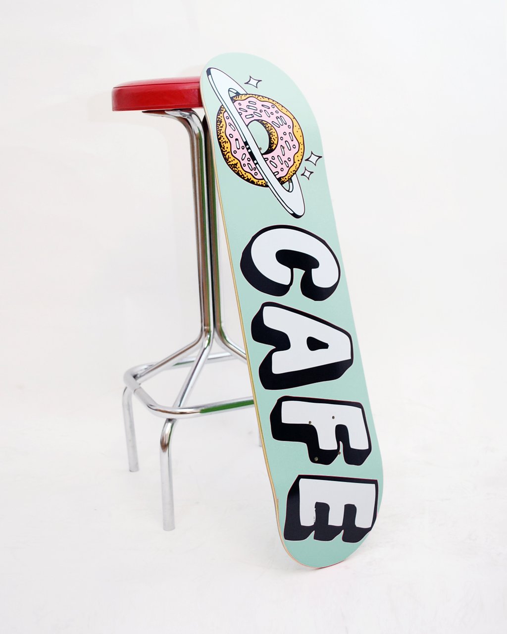 Skateboard Cafe Planet Donut Icing Blue skateboard deck: Pink-iced sprinkled doughnut ringed like a planet with sparkles above large white block letters reading CAFE with black drop shadows, on a pale mint deck.
