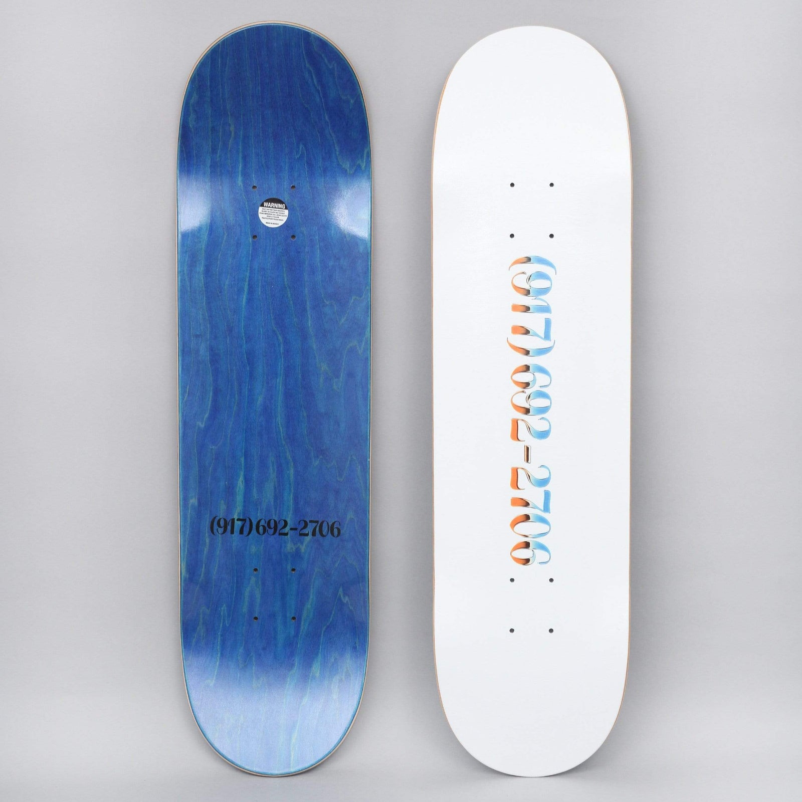917 17 8.25 Chrome Dialtone Slick White skateboard deck: A white deck with a phone number set vertically in ornate chrome-effect numerals shaded blue and orange. The blue-stained underside repeats the number in plain black type with a warning sticker.
