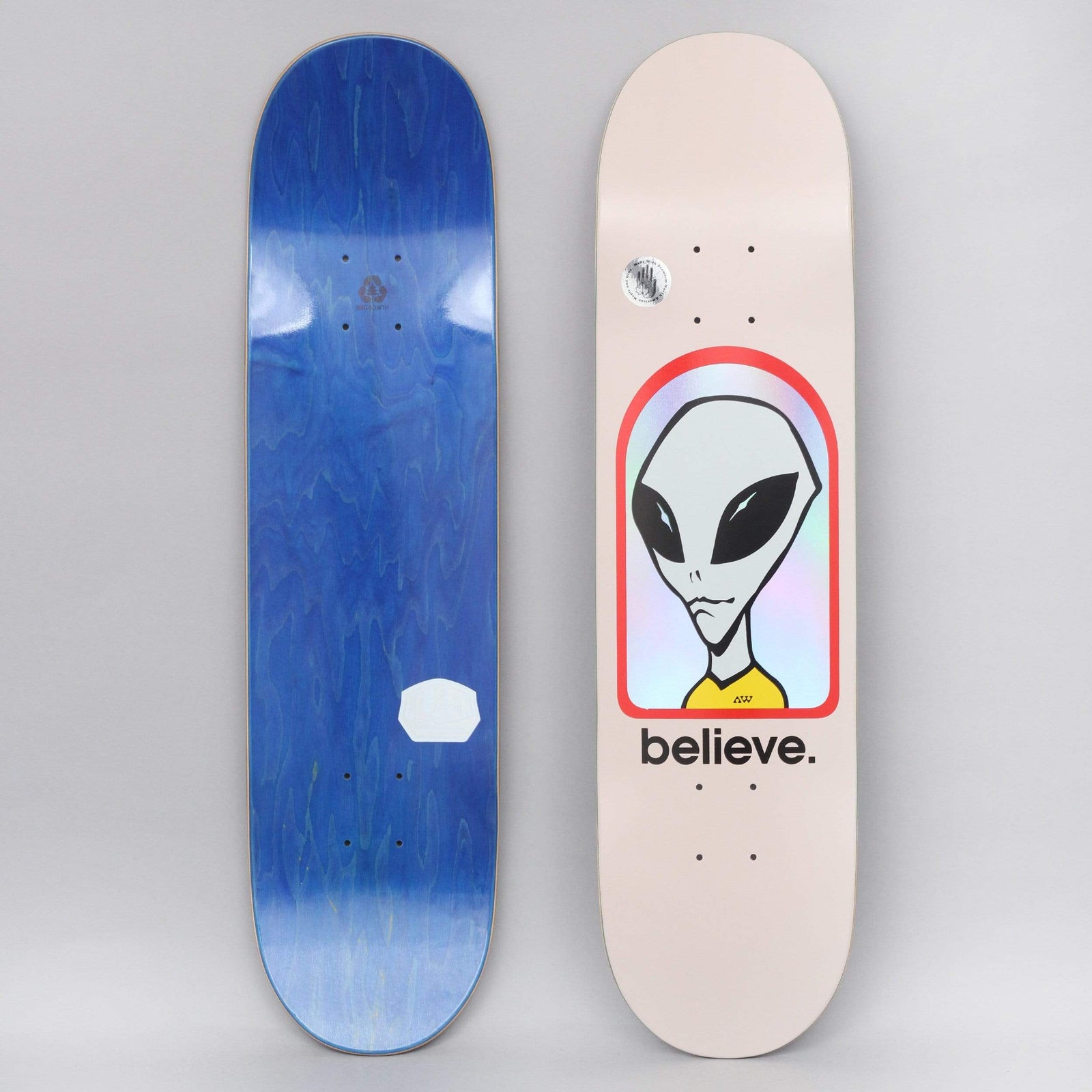 Alien Workshop Believe Hologram Pink skateboard deck: Pale pink deck with a grey alien head and yellow collar inside a red-outlined arch filled with holographic foil, with believe. in black lowercase below. Bottom view shows a blue-stained underside.
