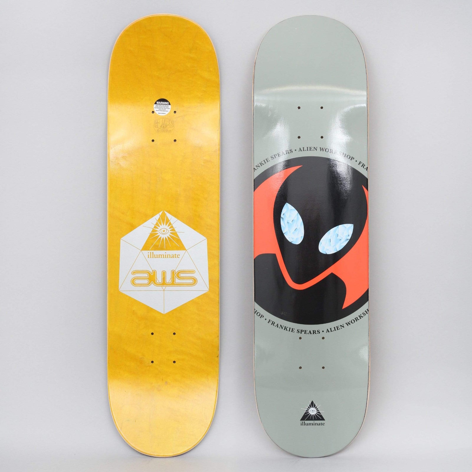 Alien Workshop Frankie Spears Sacred Geo Black / Grey skateboard deck: A large black alien head with orange edging and glittering pale blue eyes sits in a circle on a grey deck, ringed by repeated 'Frankie Spears Alien Workshop' text. The photo also shows the underside: yellow wood with an 'aws illuminate' hexagon and eye-in-triangle symbol.