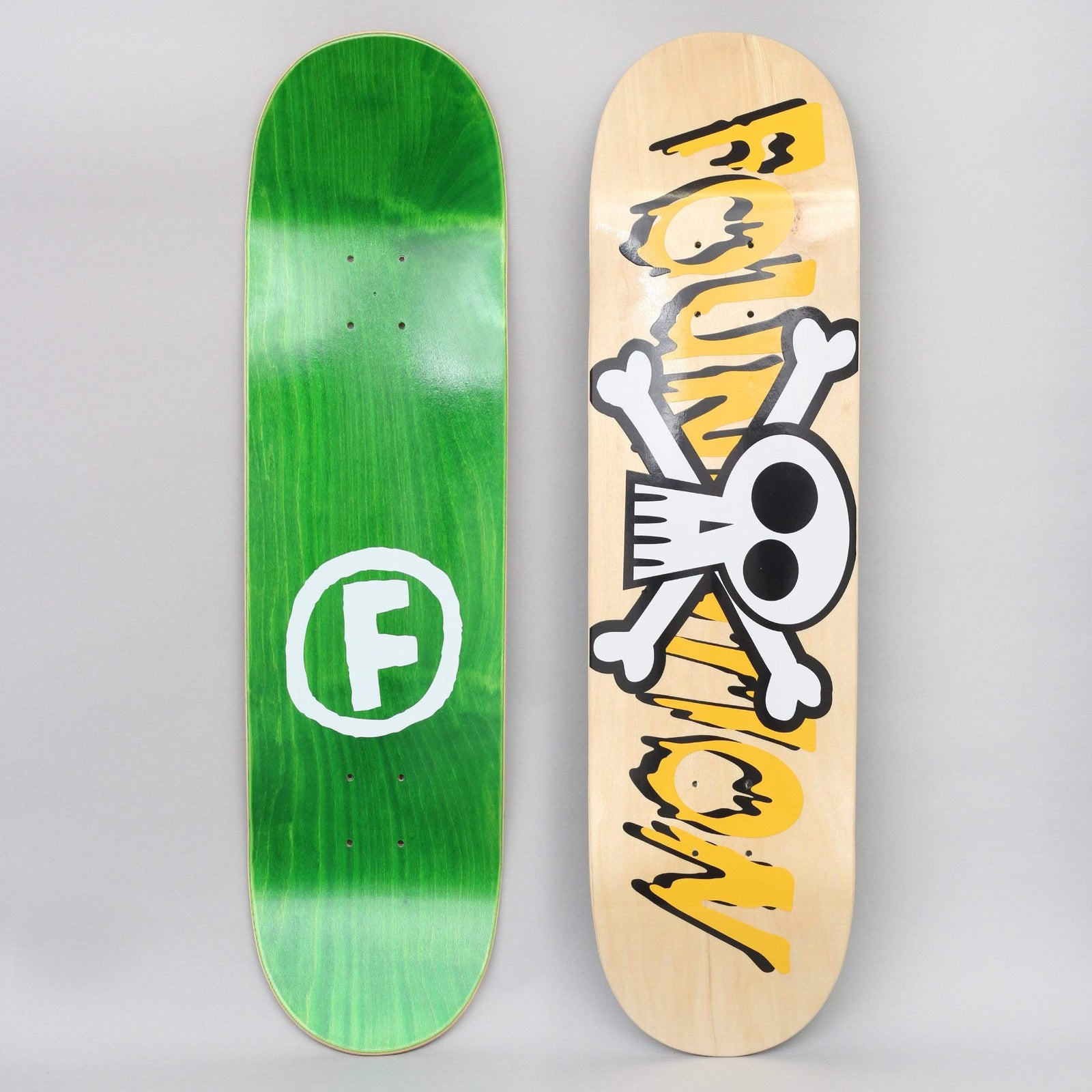 Foundation Crossbones Natural / Yellow skateboard deck: A cartoon white skull and crossbones overlaps large dripping, melting 'Foundation' lettering in yellow and black on a natural wood deck. The photo also shows the green-stained top with a white circled F logo.