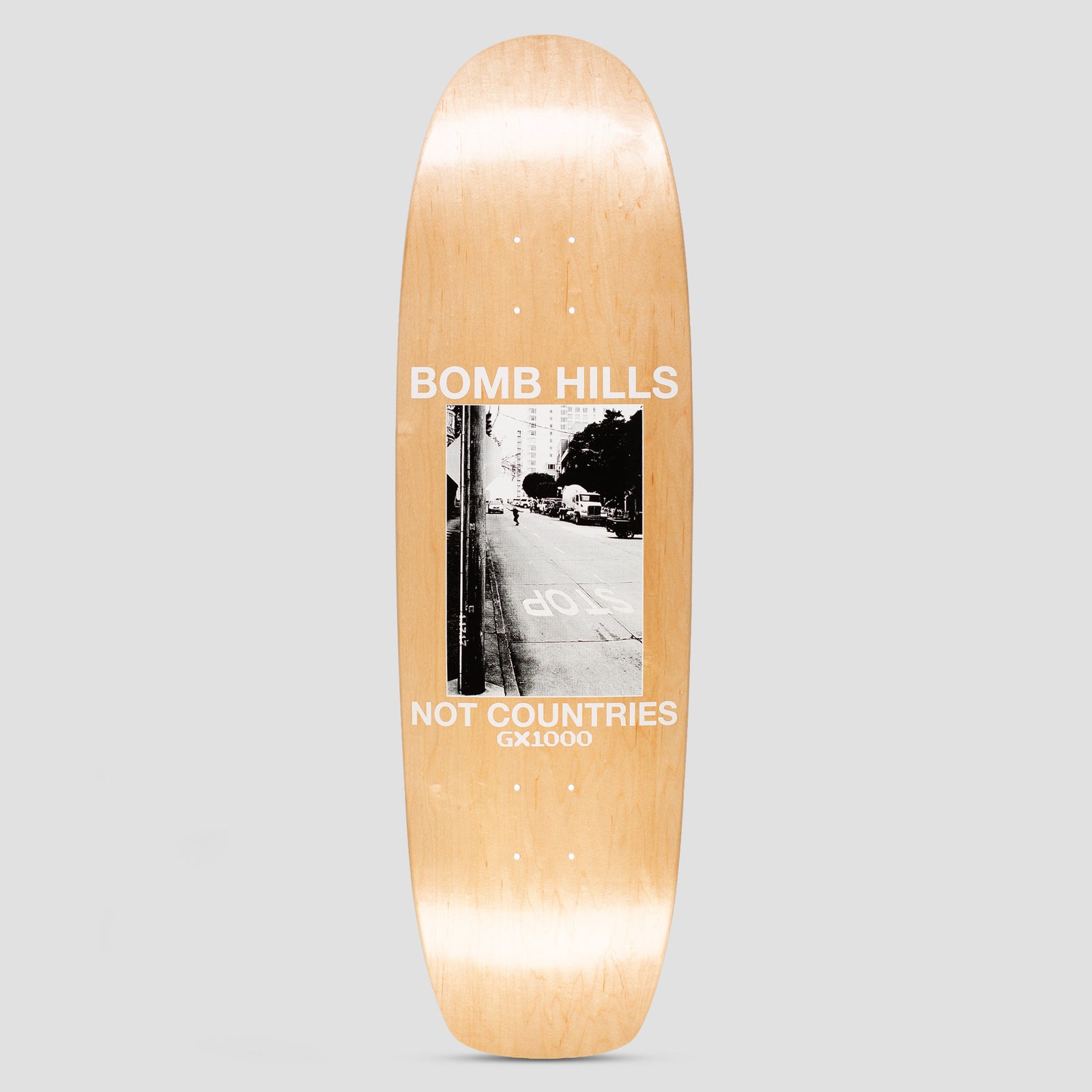 GX1000 Bomb Hills Cruiser skateboard deck: Black and white photograph of a city street with a STOP marking on the road and a skater in the distance, in a framed panel between 'BOMB HILLS' and 'NOT COUNTRIES' in white capitals, on a natural wood-stain shaped cruiser deck. GX1000 below.