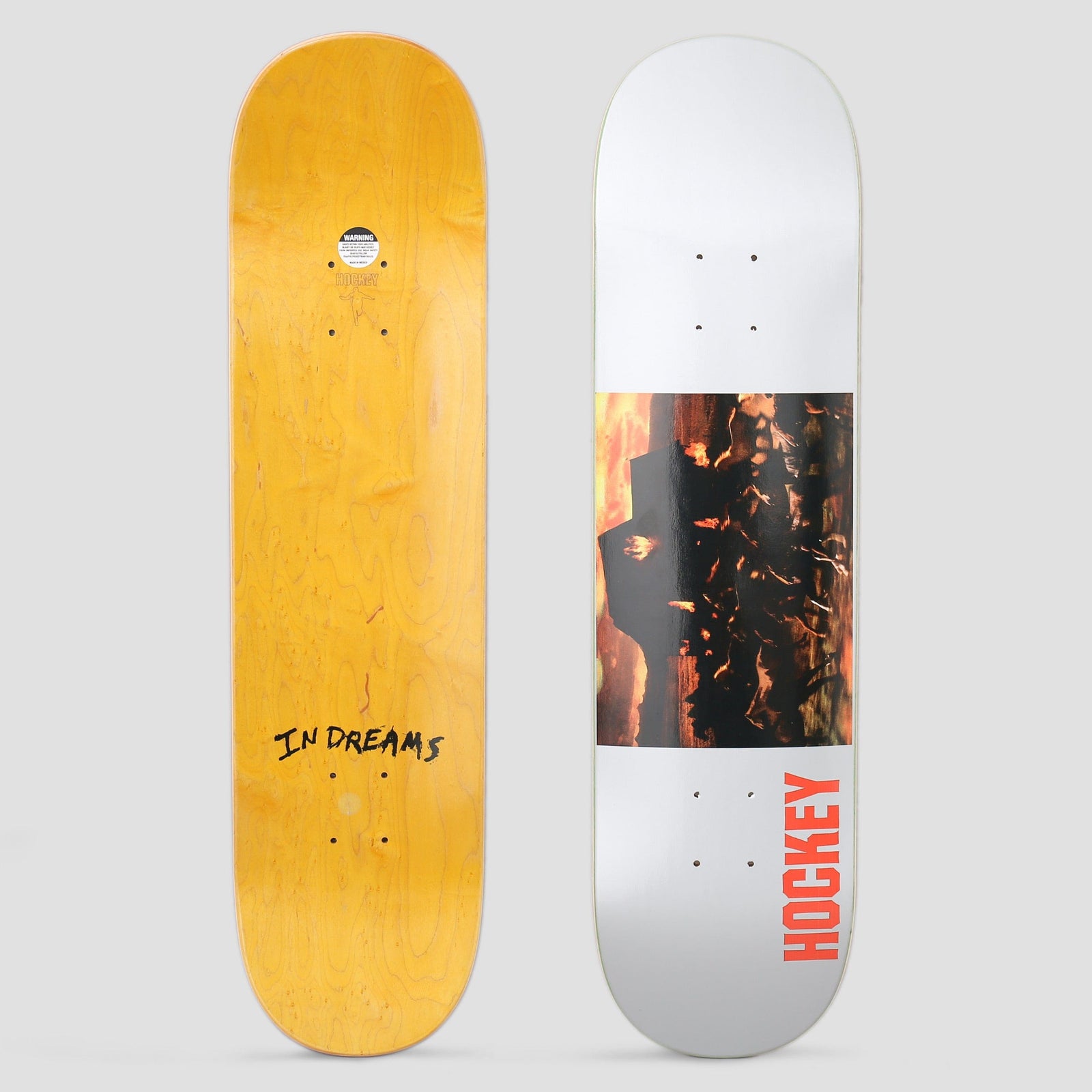 Hockey In Dream Silver skateboard deck: Dark photograph of a burning, fiery landscape with a silhouetted shape against an orange sky, set as a band on a silvery-white deck with red Hockey lettering running up the lower edge.