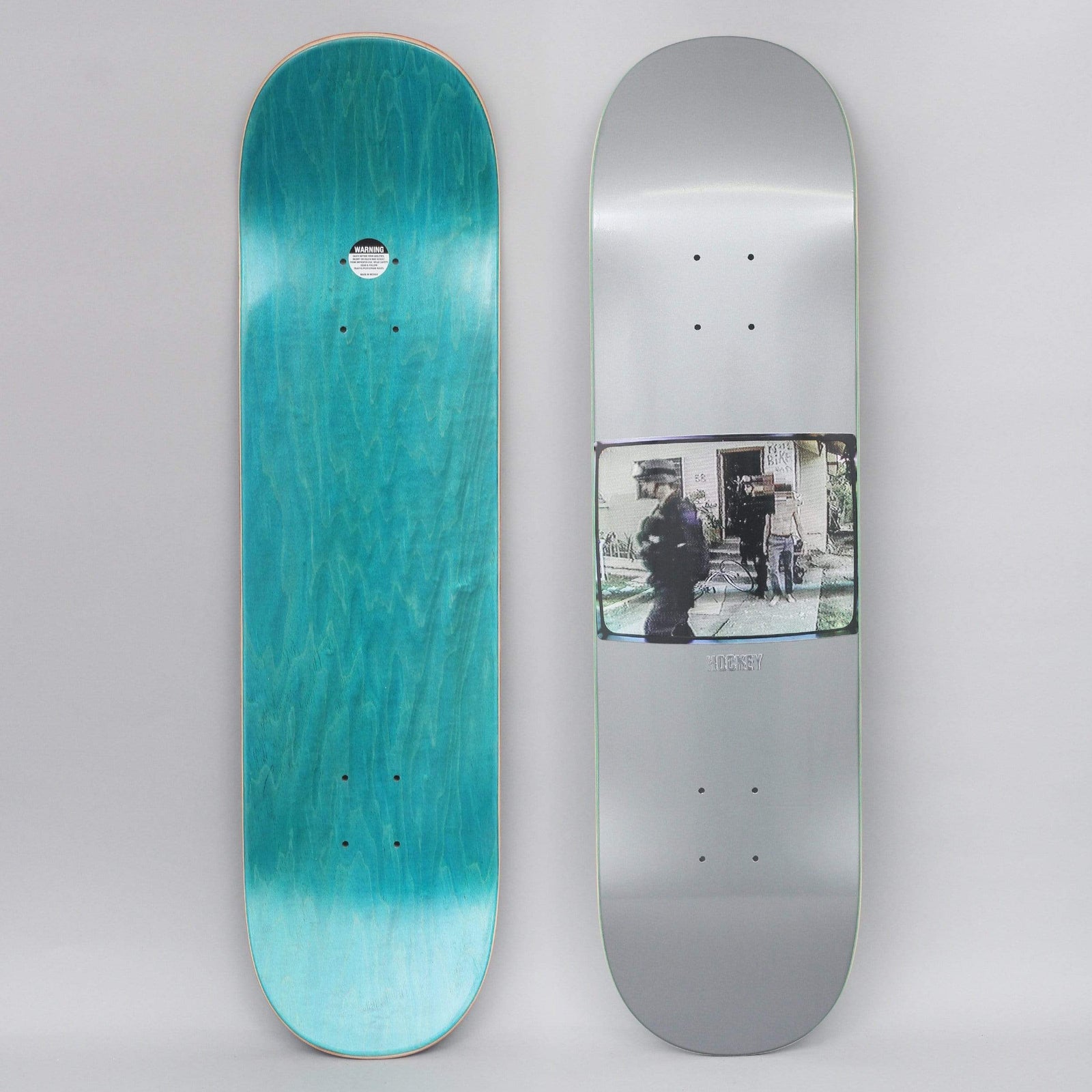 Hockey Ricks Silver skateboard deck: Metallic silver deck with a small rectangular grainy photo in the lower middle showing figures in dark clothing outside a house, with faces obscured. Small Hockey text beneath it; teal stained bottom view alongside.