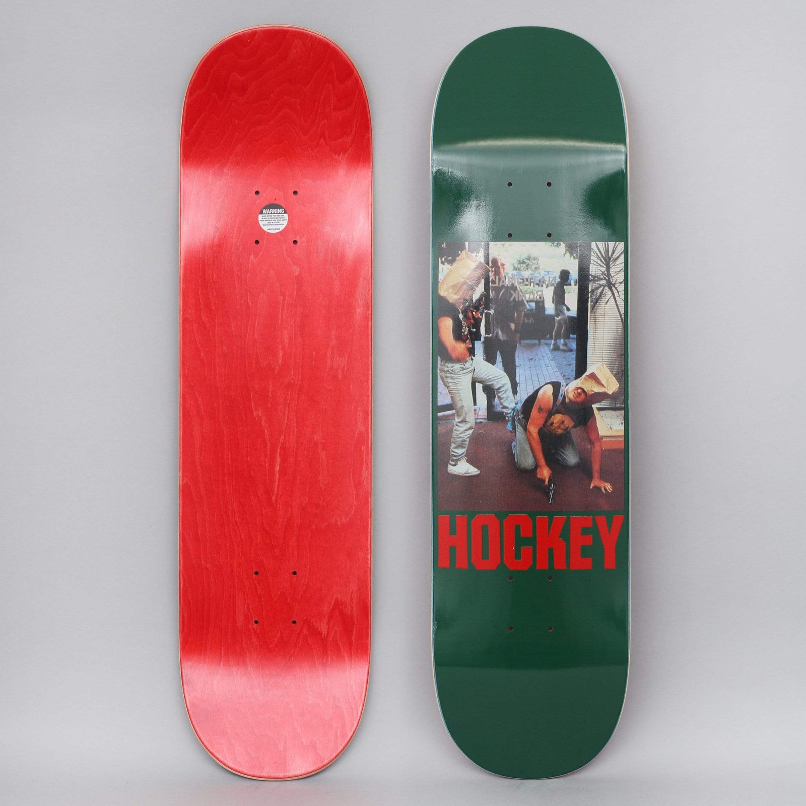 Hockey Baghead 2 Green skateboard deck: Colour photograph of two men with paper bags over their heads, one standing and one crouching with a pistol, on a street outside a shop. Red HOCKEY lettering below on a dark green deck.