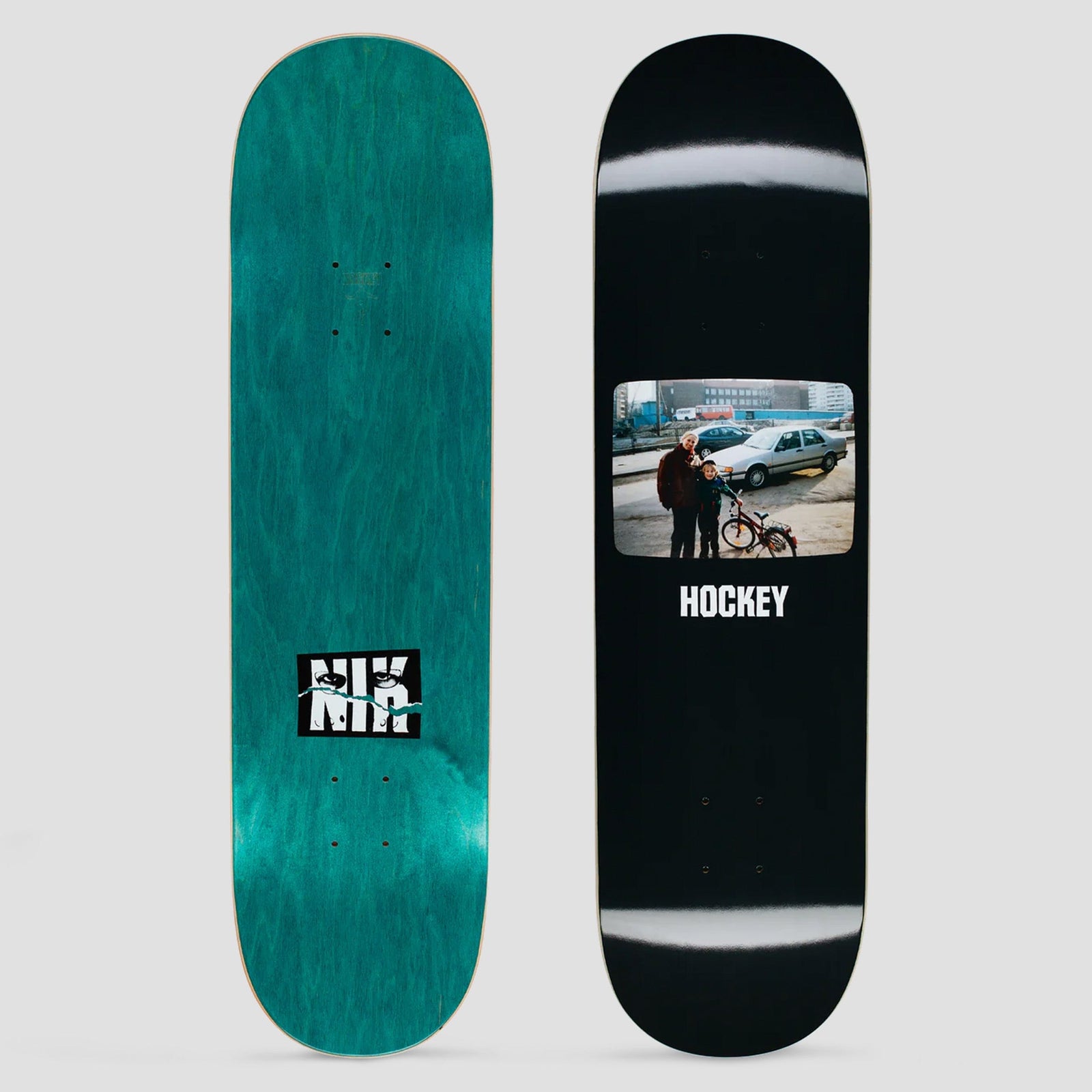 Hockey Irina Nik Stain skateboard deck: Rectangular colour snapshot of two people standing by a child's bicycle and a silver car on a bare street with tower blocks behind, above the brand name on a black deck. Teal underside has a black and white Nik logo.