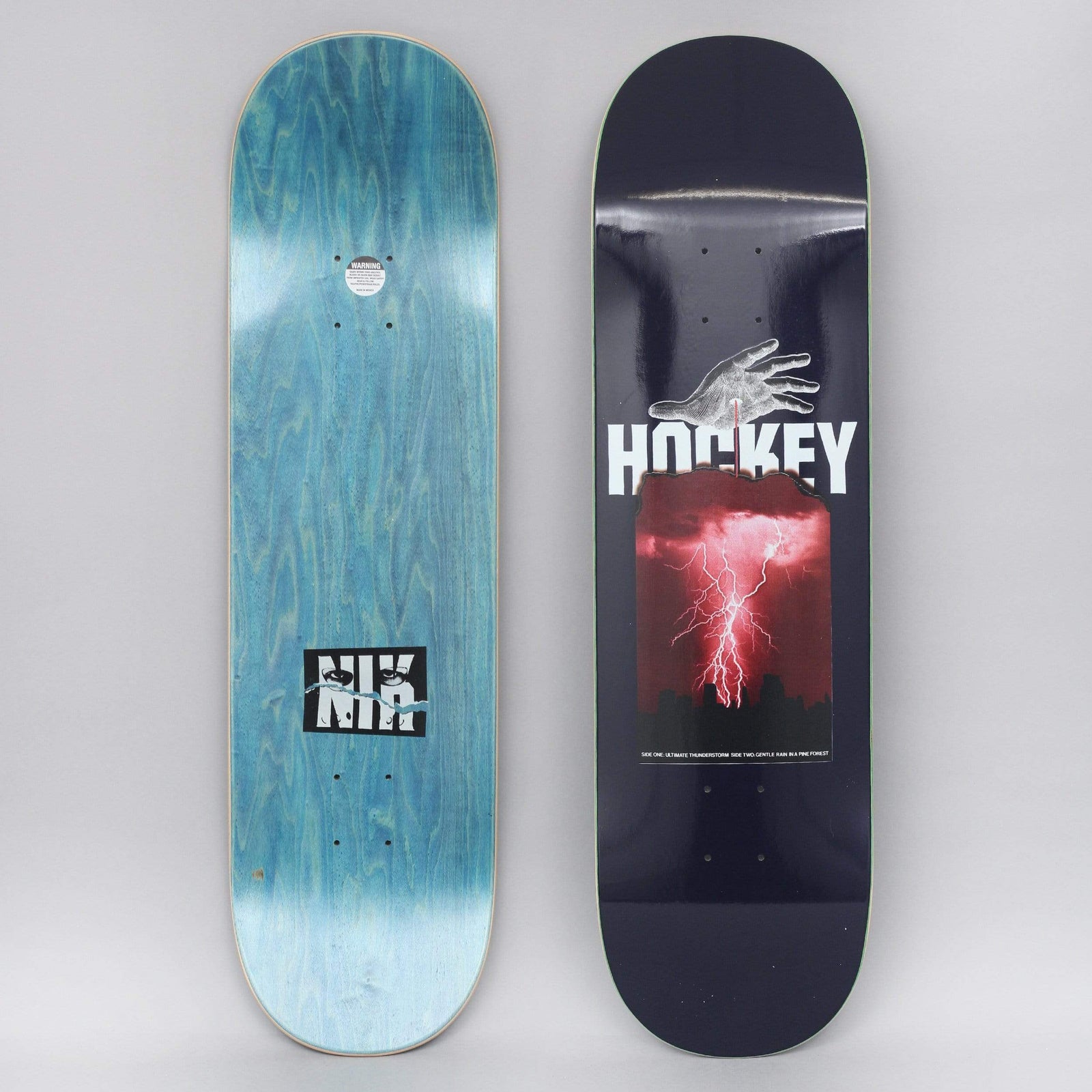 Hockey Stain Side Two Navy skateboard deck: Navy deck with a pale hand reaching down towards a bold white Hockey logo, over a red-tinted photo of a lightning storm above a city skyline. Small caption text beneath; the bottom shows a blue-stained deck with an NK logo.