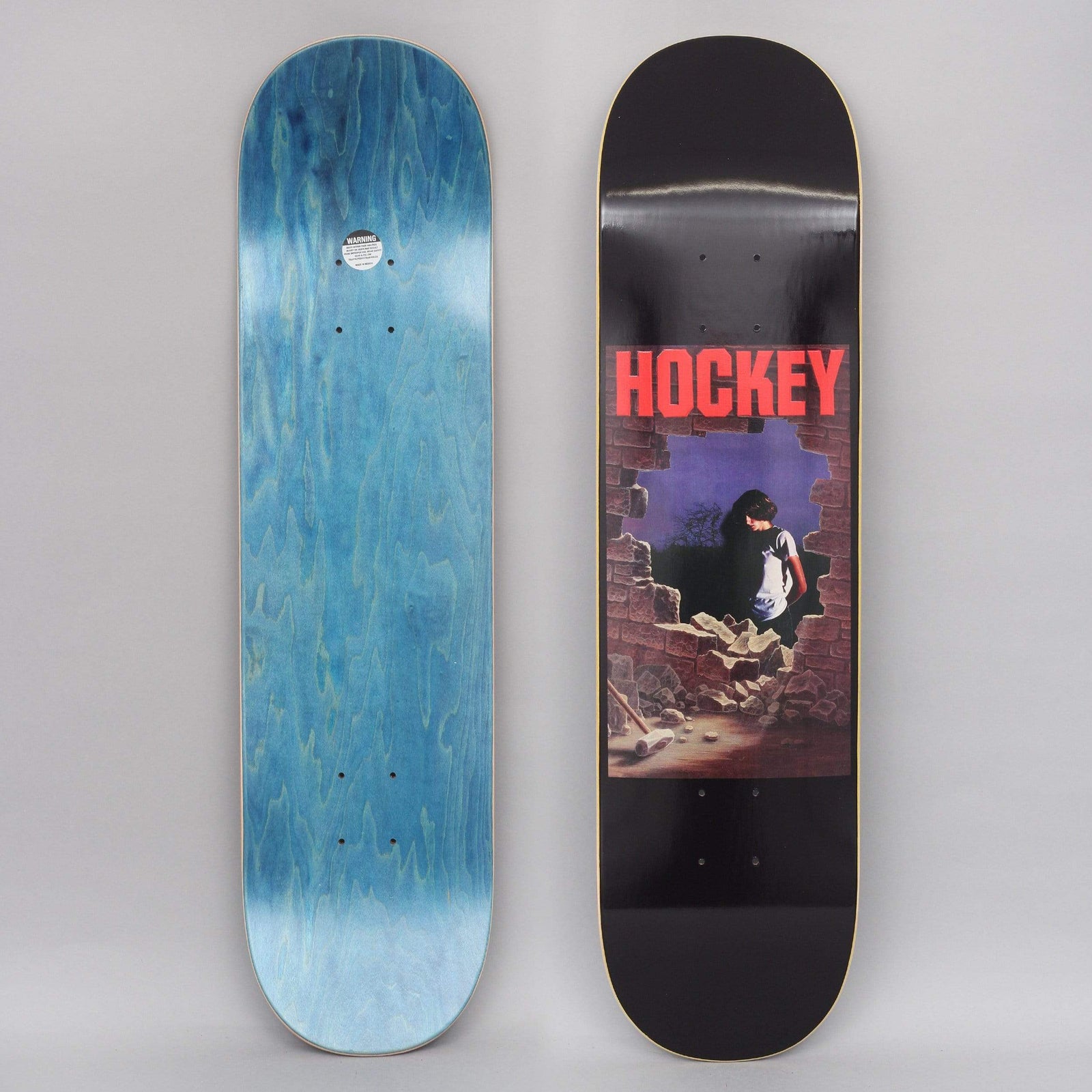Hockey Donovon Piscopo Dawn Black skateboard deck: Painted panel of a dark-haired person in a white t-shirt standing in a hole smashed through a brick wall, with rubble and a sledgehammer on the floor. A red Hockey logo sits above, on black.