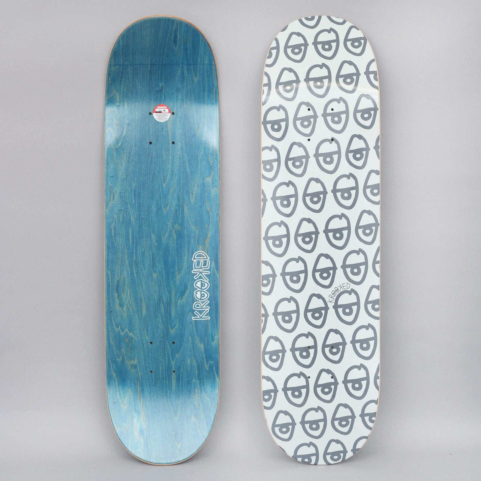 Krooked Pewpils Grey skateboard deck: The graphic is a repeating all-over pattern of grey cartoon eyes on a pale ground. A deck with blue wood-veneer underside is shown beside it.