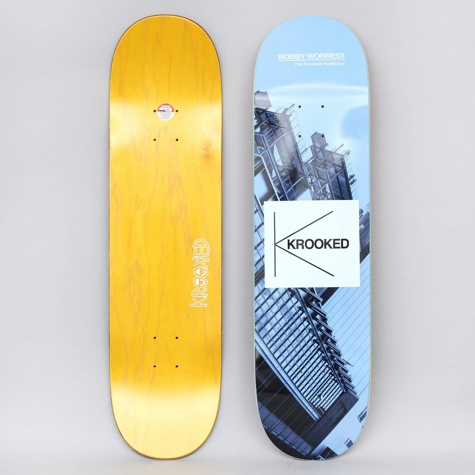 Krooked Worrest Kollection Full Blue skateboard deck: Blue-tinted photograph of a modern glass and steel building on a tilted angle, with a white square carrying the Krooked K logo. Rider name and 'The Krooked Kollection' are printed at the top. Photo shows both the graphic and the yellow bottom.