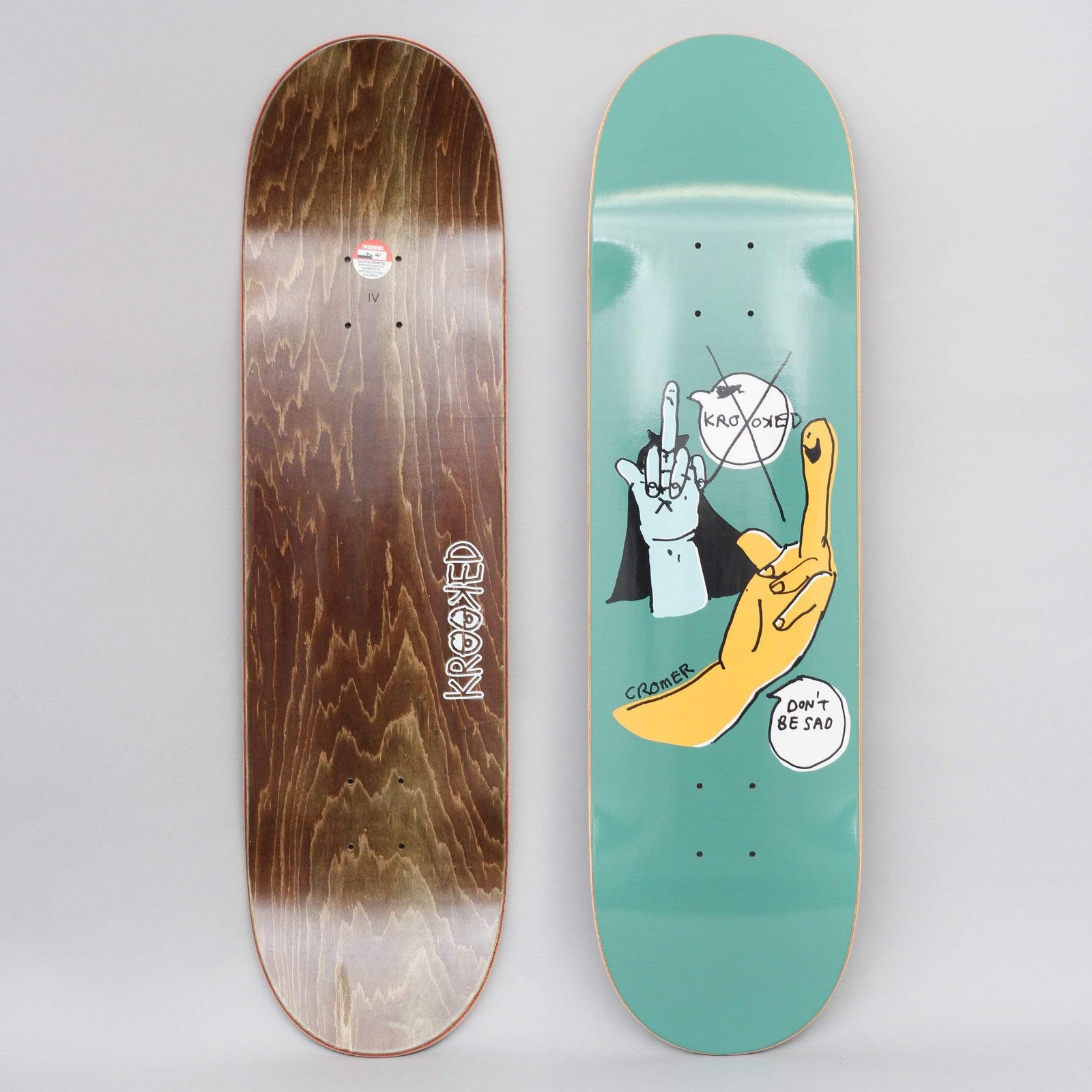 Krooked Cromer Redux Green skateboard deck: Two hand-drawn hands making rude gestures, a blue vampire-cuffed one and a larger yellow one with a smile, on a green-teal deck with speech bubbles. Brown wood underside shown alongside.