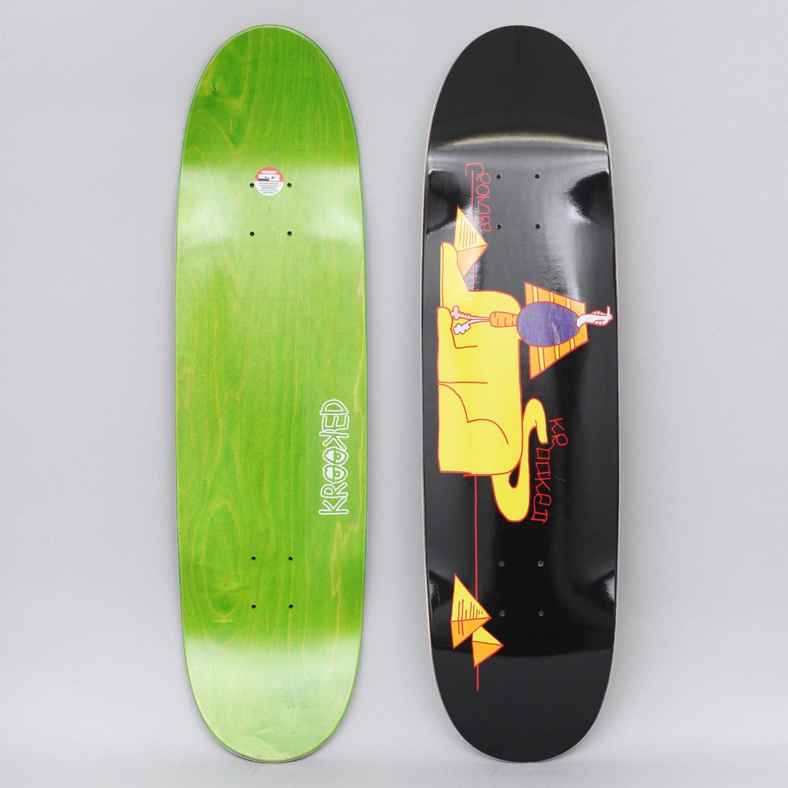Krooked Cromer Sfinkgx Black skateboard deck: Black shaped deck with a yellow reclining sphinx-like figure, a small purple-bodied figure standing on it, and orange pyramids with a red line. Photo also shows the green-stained underside with Krooked logo.