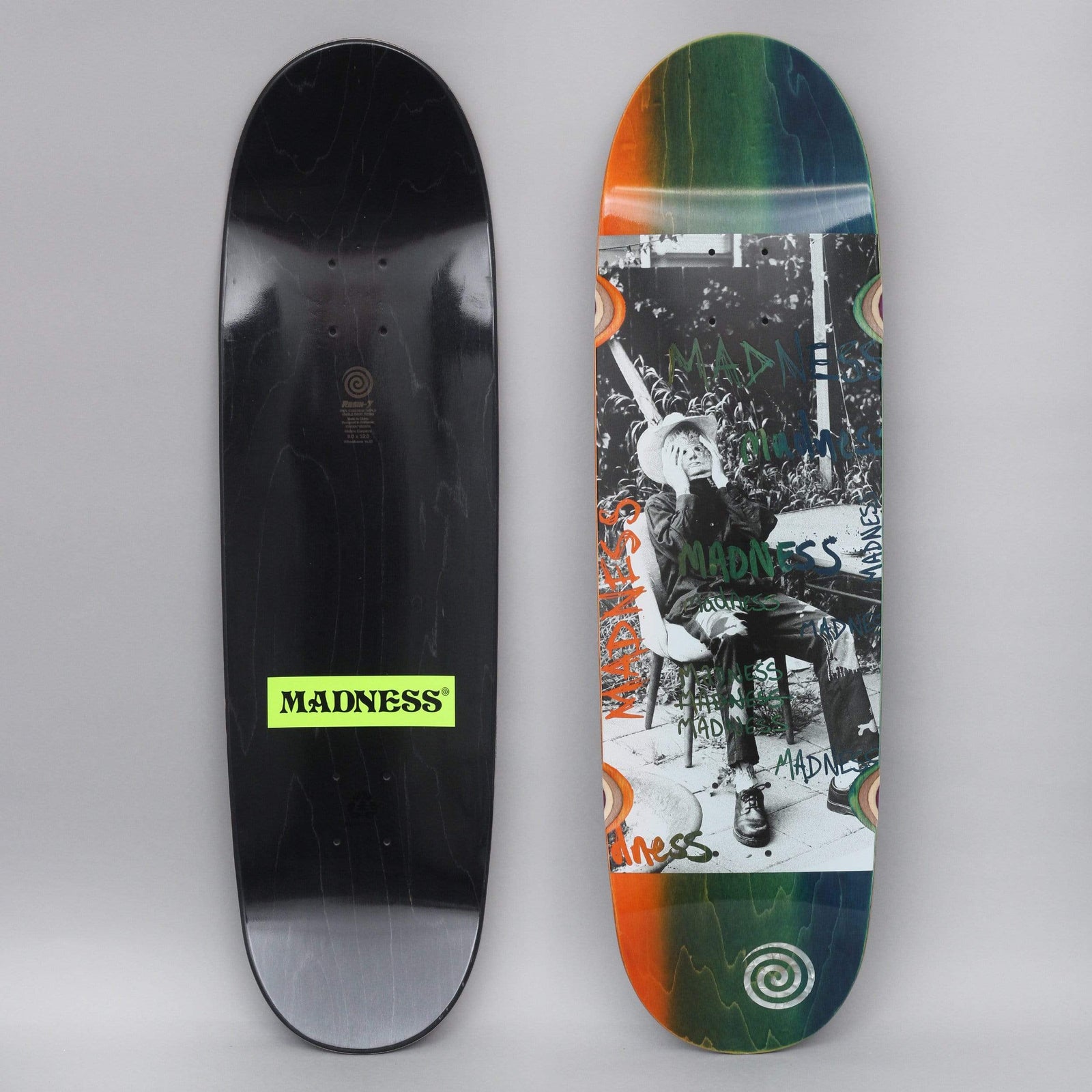 Madness Column R7 Blue / Orange skateboard deck: A black-and-white photo of a seated person in a hat covering their face with their hands, overprinted with green and blue hand-drawn Madness scrawls, on an orange-to-green-to-blue faded deck with spiral logos. The underside is black with a green Madness label.