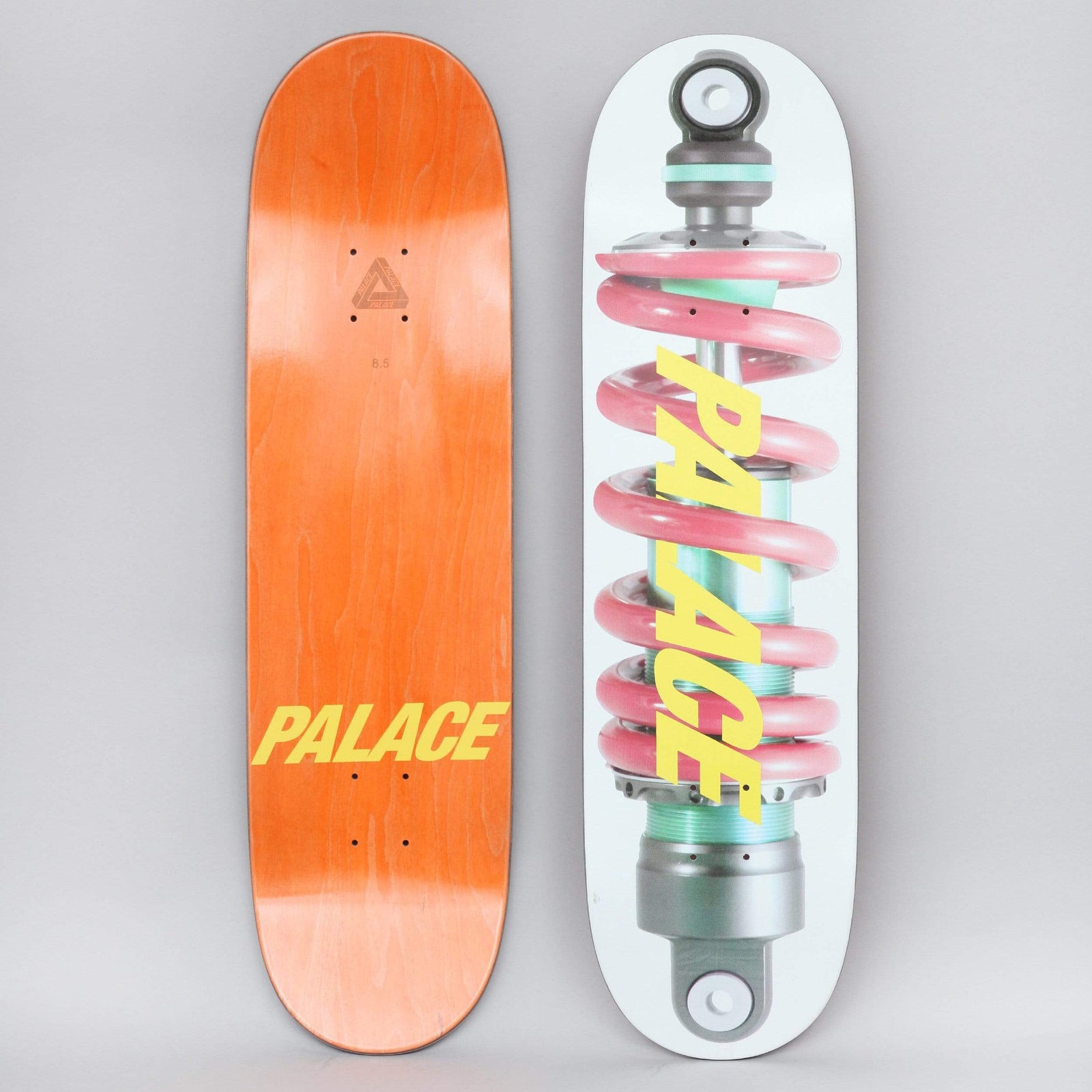 Palace Shock White / Yellow skateboard deck: A close-up photographic image of a motorbike or car shock absorber with a thick pink coil spring and mint green body, on a white deck. The Palace name is printed diagonally in yellow across the spring; the orange underside is also shown.
