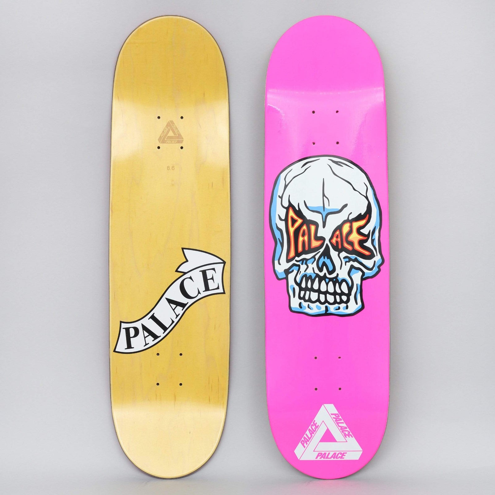 Palace Hesh Pink skateboard deck: Cartoon white skull with blue shading and the word Palace in orange flame-style lettering across its eye sockets, on a bright pink deck with a white Palace tri-ferg logo near the tail.