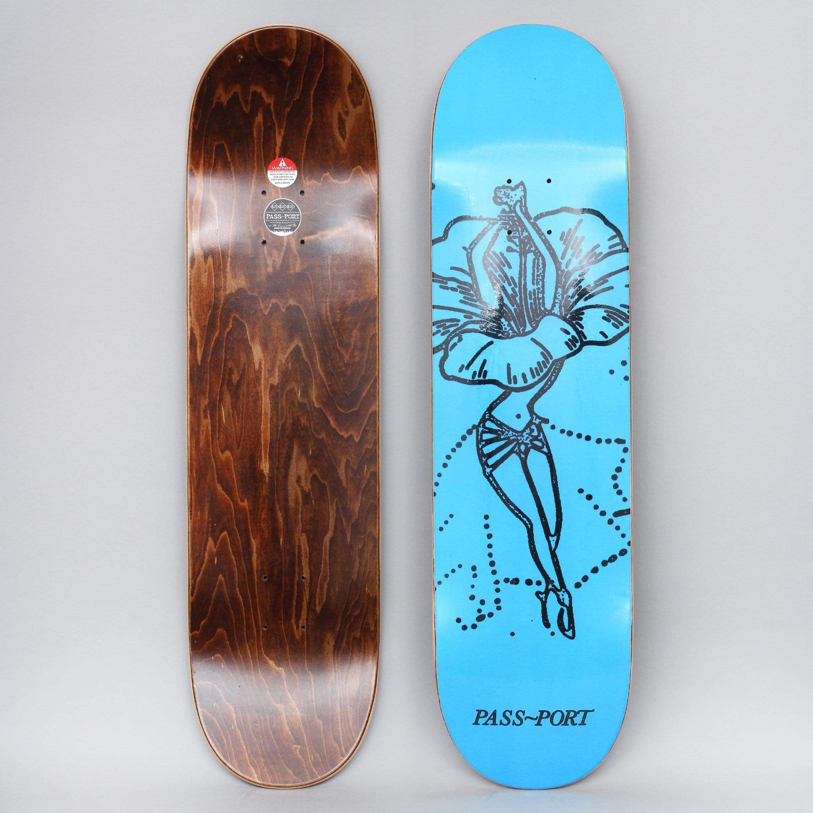 Passport Floral Dancer Hibiscus Blue skateboard deck: Black line drawing of a dancer with a hibiscus flower as her skirt, standing on pointe with dotted arcs around her, on a bright blue deck. Brand name in serif capitals at the foot. Brown wood underside shown beside it.
