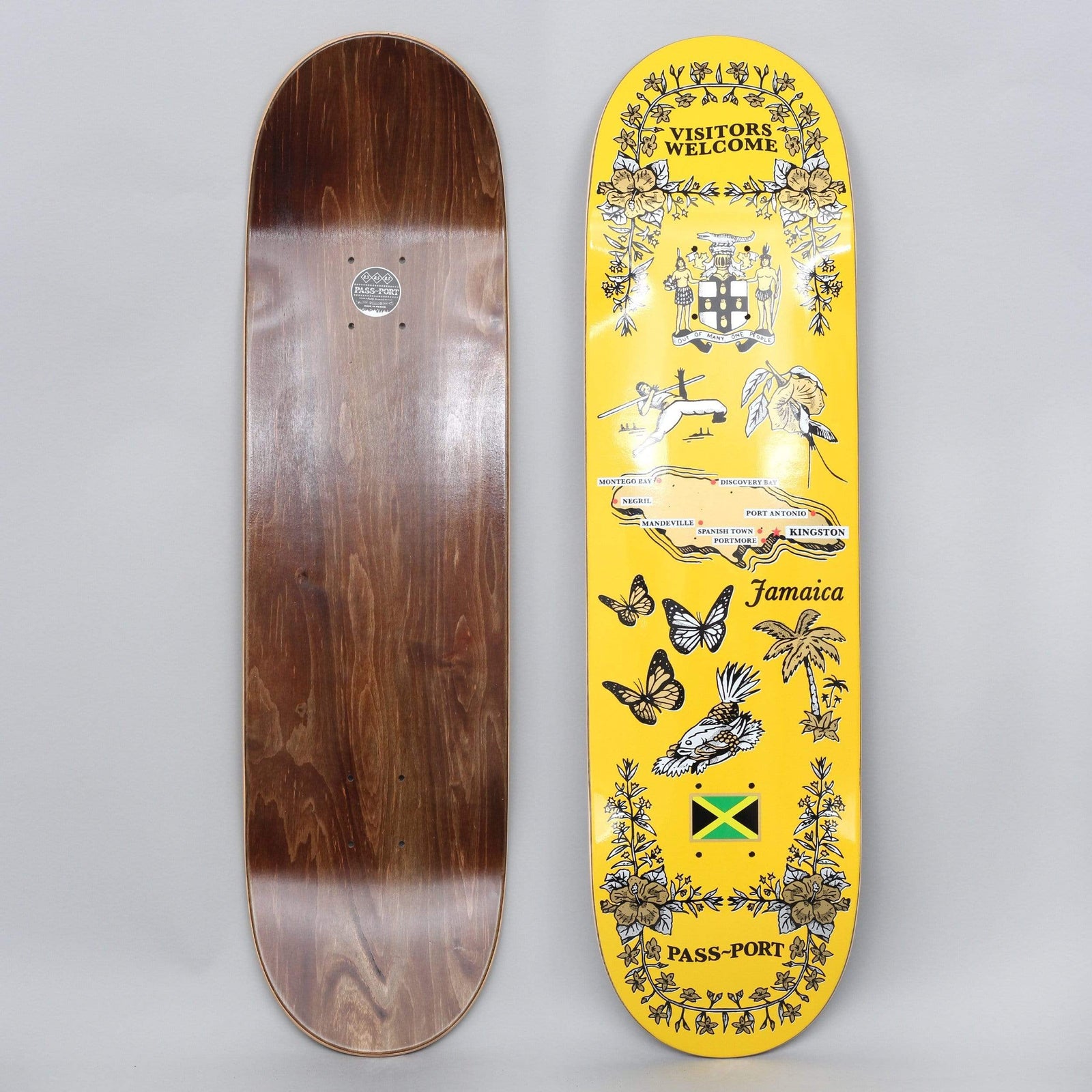 Passport Tea Towel Jamaica Yellow skateboard deck: Yellow deck styled as a Jamaica souvenir tea towel: floral borders, coat of arms, a map with town names, butterflies, a bird, a palm tree and the Jamaican flag. The other deck shows a brown stained underside.