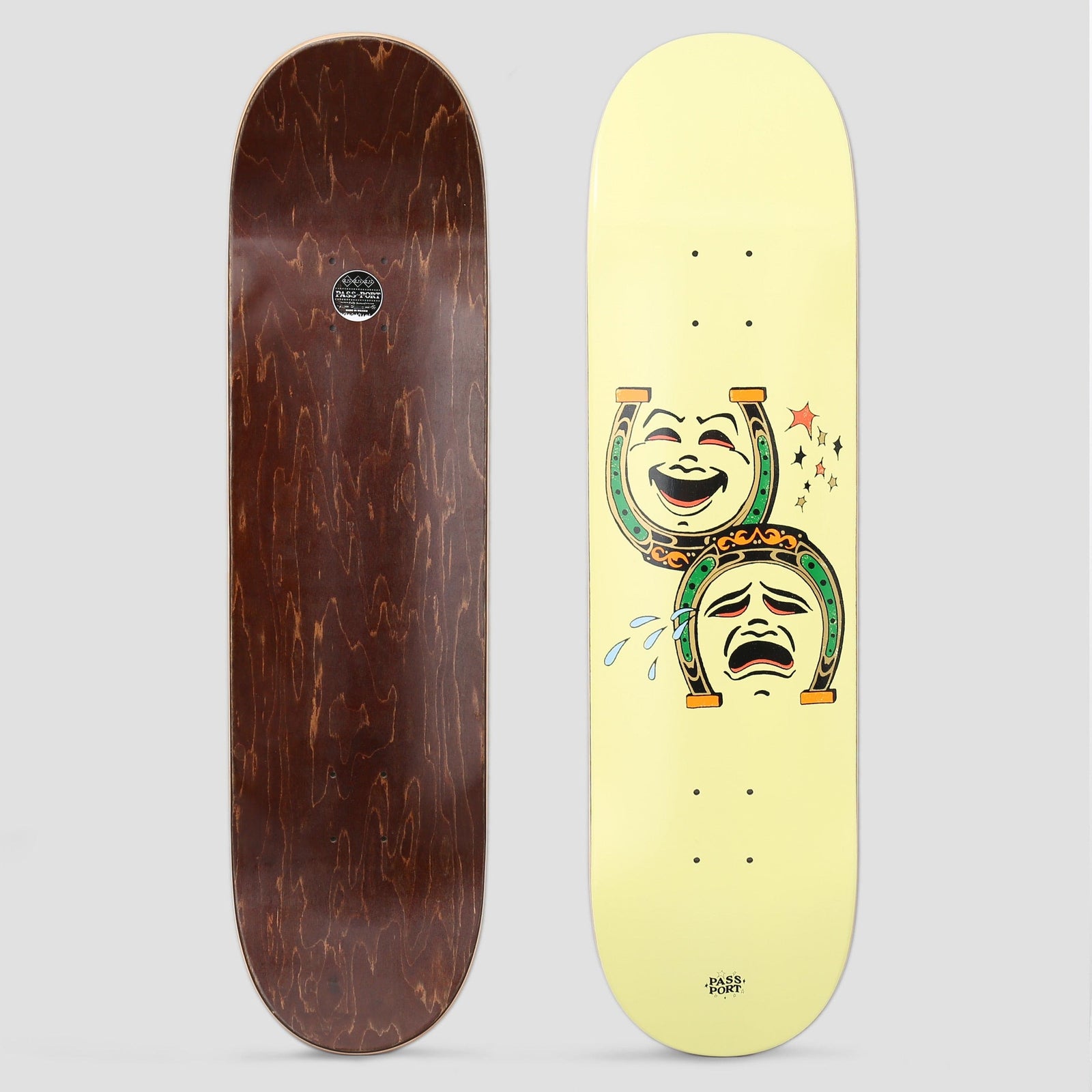 Passport Dumb Luck Series Horse Shoe Yellow skateboard deck: Two linked horseshoes in a tattoo-flash style, one framing a laughing comedy face and the other a crying tragedy face with tears flying off, plus small stars, centred on a pale yellow board.