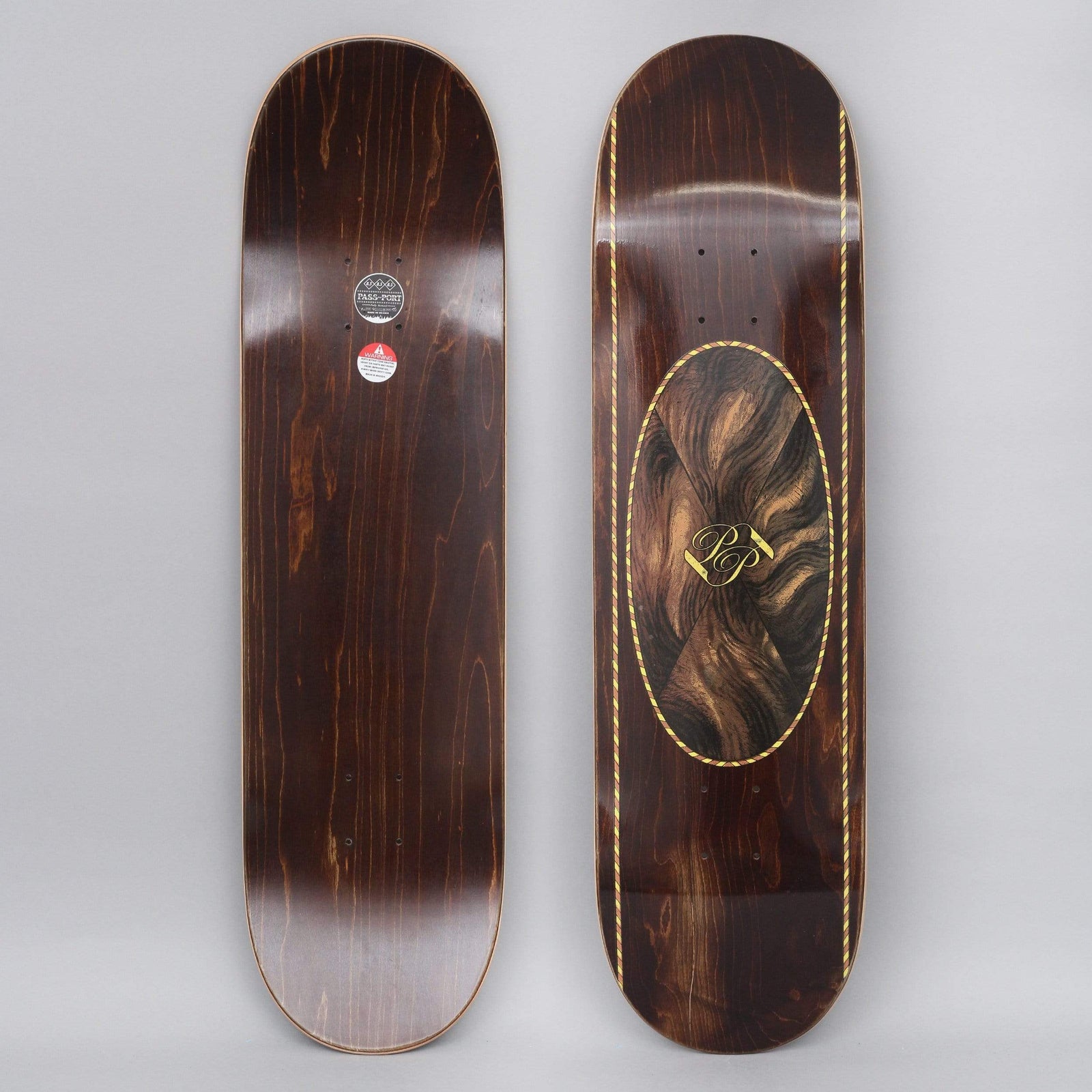 Passport Inlay Brown skateboard deck: Dark brown wood-stained deck with an oval panel of swirling wood-grain veneer pattern and gold PP monogram, edged with a gold rope-style border running down both sides.