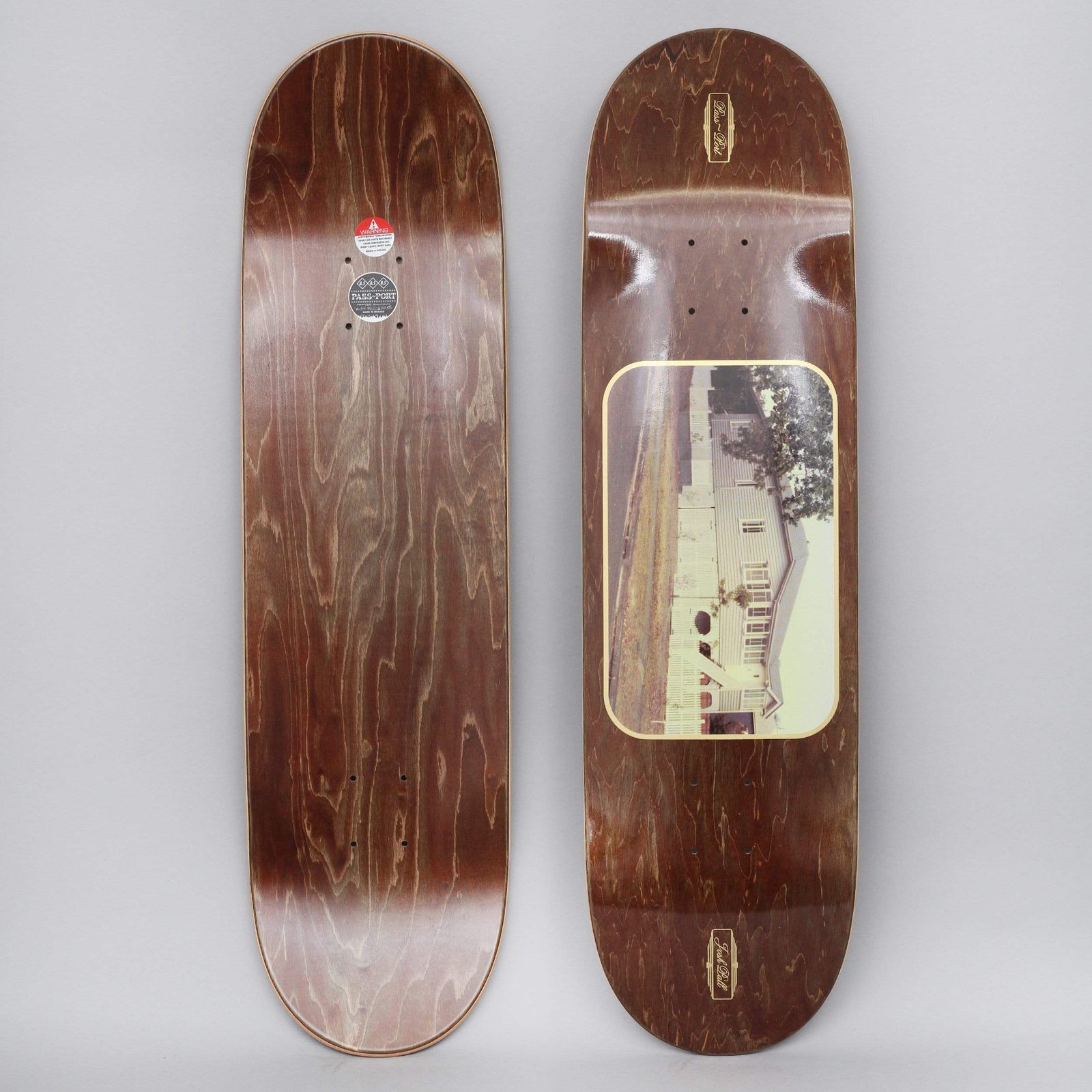Passport Josh Pall Home skateboard deck: Brown-stained deck with a rounded gold-bordered photo panel of a white wooden house with a tree, and small gold Pass-Port logos at nose and tail. Plain stained underside shown alongside.